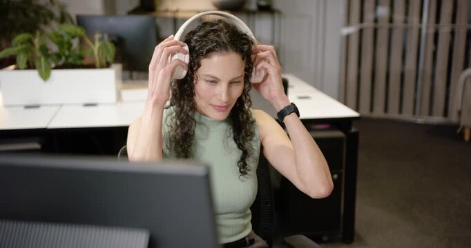 African American woman sitting at office desk reaching for white headphones while refocusing on PC
