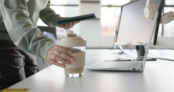 African American nonbinary walking to desk placing travel cup tapping lid organizing with tablet