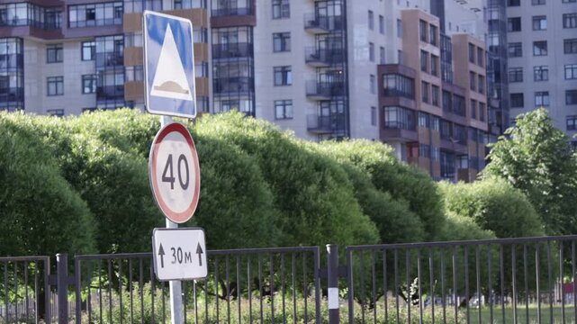 Stacked traffic signs indicate a 40 km/h speed limit with 30-meter zone extension; set against dense green shrubbery and modern apartment buildings, the scene reflects a regulated urban street environ