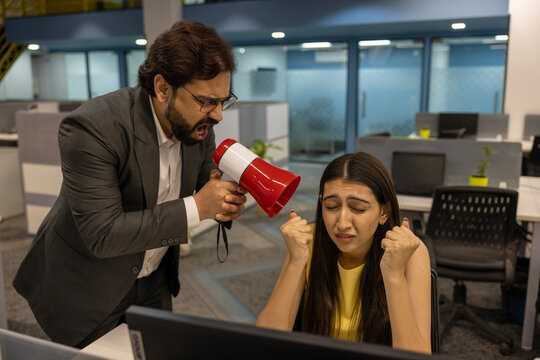 Indian angry businessman shouting at employee woman holding megaphone standing in modern office. professional communication problem sitting at the desk who does not want to work.
