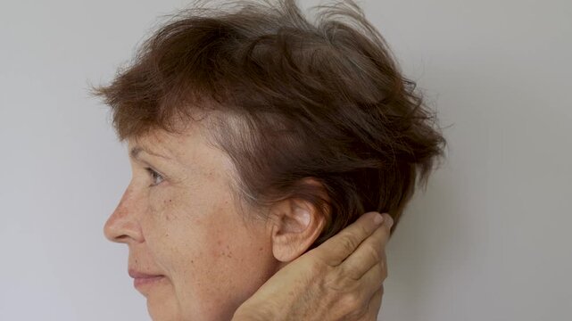 Mature woman lifts hair at the temple, showing gray roots and natural graying close-up on a light background