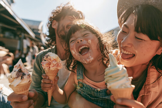 Happy Asian family enjoying ice cream outdoors, young girl with messy face laughing between parents, sunlight, summer day, joyful, candid, bonding moment