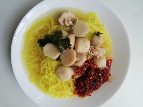 Top view of yellow rice vermicelli noodle soup served with fishballs, chopped Chinese celery, and spicy chili oil on a white plate. 