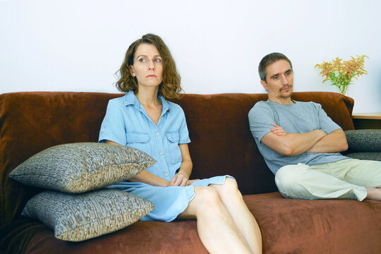 Woman with tense expression looks away while man behind her sits with arms crossed on brown sofa. Illustrates silent standoff, unresolved conflict, and emotional distance between adult partners.