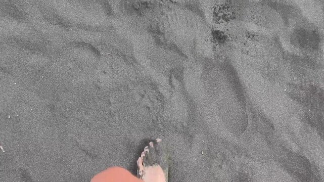 Person's bare feet walking on a volcanic black sand beach, leaving footprints in the wet, dark sand as a symbol of travel, vacation, and enjoying nature alone
