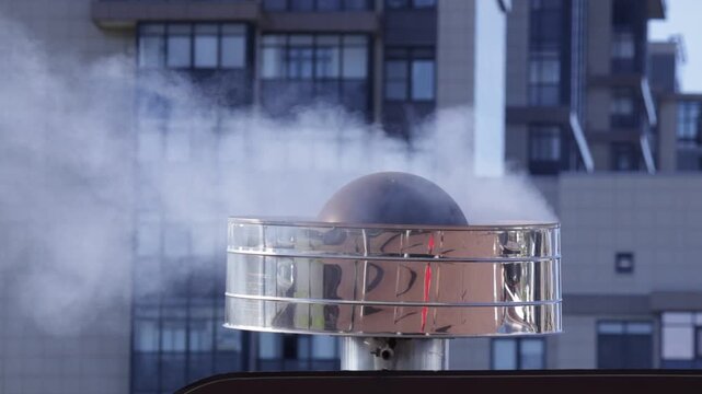 Shiny metallic vent cap atop building releases thick steam plume; urban backdrop with modern windows creates cool industrial mood