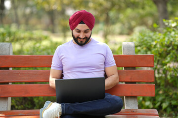 Fototapeta premium Young Indian Sikh man working on laptop while sitting on bench at summer park.