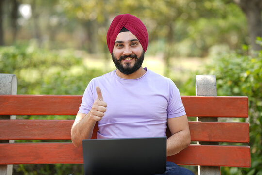 Portrait of cheerful happy sikh man showing thumbs up while working on laptop with sitting on bench at park.