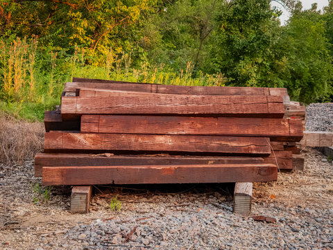 A stack of weathered wooden railway ties or sleepers sitting on gravel ballast near a green forest edge outdoors