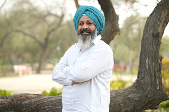 Portrait of smiling senior Punjabi bearded sikh man wearing blue pagdi and traditional white kurta and pajama outfit standing cross arms at park and looking Infront of the camera.