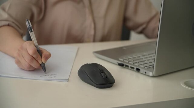 Close up of a woman writing with a pen on paper beside a laptop and wireless mouse at an office desk. Everyday note taking, planning and office work.