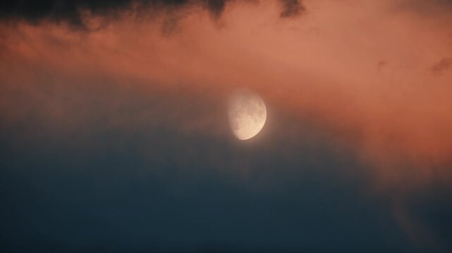 The moon behind moving illuminated orange clouds in the dark blue sky at sunset. Footage of the lunar view at dusk. 