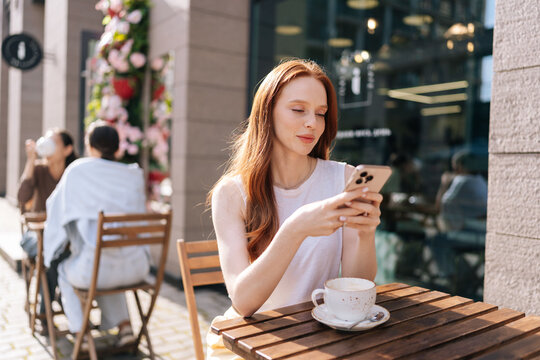 Attractive young woman with long red hair sitting at outdoor cafe table on sunny day. Redhead female using mobile phone enjoying cup of coffee. Relaxed urban lifestyle scene with blurred background.
