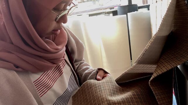 Young Asian Muslim woman in hijab examining and choosing fabric swatches and color samples for interior design project in a shop.