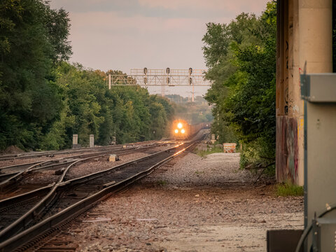 Freight train approaching with glowing headlight along parallel railroad tracks through a scenic wooded landscape