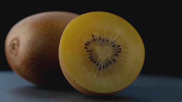Macro shot of a ripe golden kiwi cut in half with a heart-shaped core, rich juicy pulp and seeds on a dark background with a blurred whole kiwi for healthy eating, gourmet food, and culinary content.