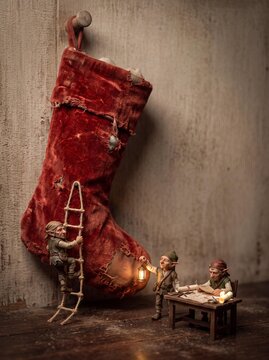 Miniature elves working around a vintage red Christmas stocking with a lantern and ladder, fantasy holiday diorama with large copy space
