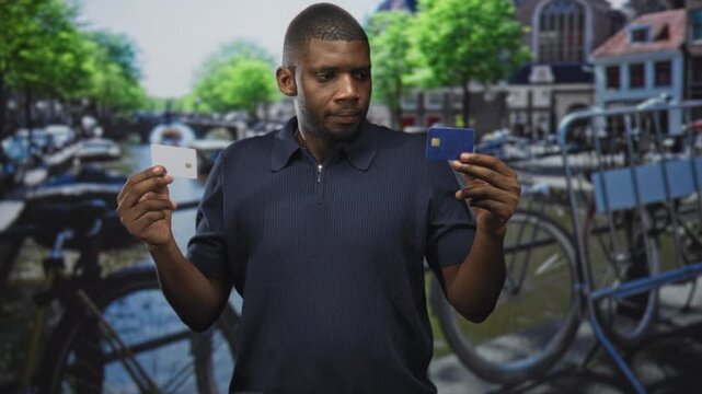 Man holding two creditcard, comparing a blue card and a white chip card with a puzzled look on a canal street; doubt decision.
