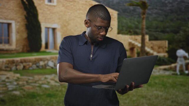 Young man typing on a laptop, holding it with one hand in front of a stone building; focus productivity.