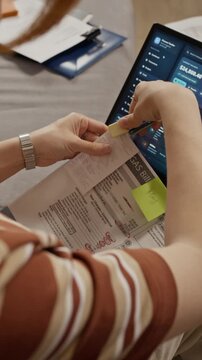 Vertical shot of young Caucasian woman checking utility gas bill and typing on laptop with financial software. Perfect for personal finance app campaigns and budgeting tutorials