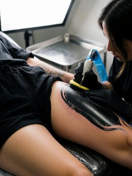 Female tattoo artist inking blackwork tattoo on client's thigh