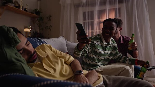 Multiethnic young adult men photographing sleeping friend with smartphone at home. Footage ideal for social media app advertising, leisure entertainment campaigns and sports fandom promotions