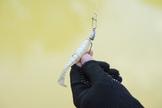 Close-up of a gloved angler holding a soft plastic fishing lure rigged with a hook, swivel and sinker, showcasing fishing tackle and artificial bait against a softly blurred outdoor background.