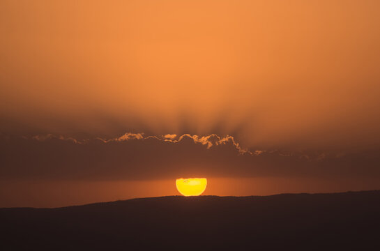 Warm sunset in the evening. The sun forms rays above a cloud and begins to hide behind a hill. Evening landscape for the background