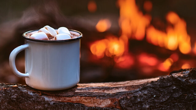 Enamel camp mug of hot chocolate with blurred autumn forest campfire background banner.
