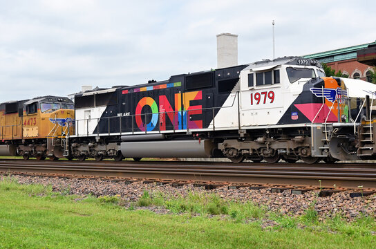Multiple Union Pacific Railroad locomotives, including one with a "We Are One" workplace equality paint scheme, lead a cotainer train eastbound through a suburban Chicago community.