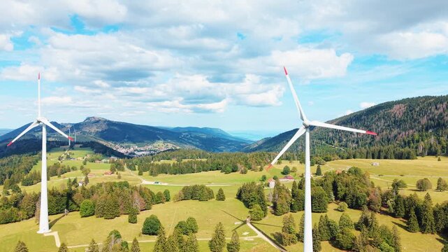 Aerial view of the Sainte-Croix wind farm featuring large turbines set against a backdrop of rolling green hills, dense forests, and a blue sky in Sainte-Croix, Vaud, Switzerland.