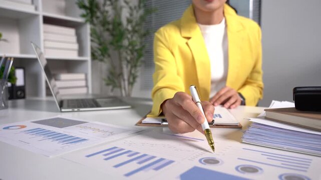 Financial Analysis: A businesswoman in a yellow blazer meticulously reviews financial reports and charts on her desk, highlighting data with a pen, in a modern office setting.