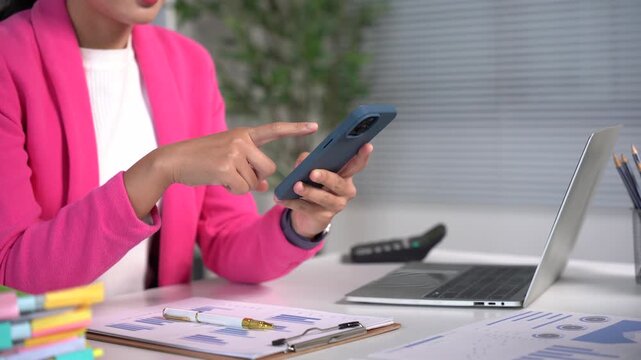Woman Using Smartphone at Desk: A professional woman in a vibrant pink blazer is seated at her desk, intently focused on her smartphone, with a laptop, calculator.