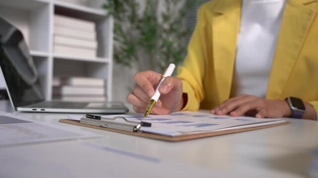 Analyzing Financial Data: A close-up shot shows a person in a bright yellow blazer meticulously reviewing financial reports and charts on a desk, with a laptop and office supplies nearby.