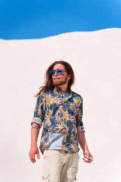 Stylish man with long brown hair, beard, and floral shirt wearing sunglasses while standing on white sand dunes against a clear blue sky during summer in Lesbos, Greece.