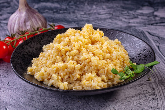 Cooked bulgur wheat in black bowl with cherry tomatoes and garlic on textured gray surface