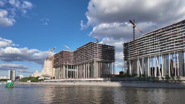 Timelapse of a residential complex under construction on slender stilts along the Moskva River embankment. Tower cranes swing over the building site. Boats and river buses pass on the water below.
