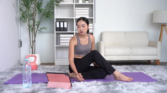 Young Woman Practicing Yoga at Home: A woman in workout attire sits on a yoga mat, following an online class on her tablet, in a modern living room.