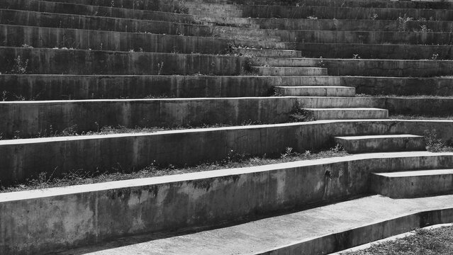 Monochrome view of weathered concrete terraced steps with repeating geometric lines, rough surfaces, overgrown vegetation, and strong architectural texture.