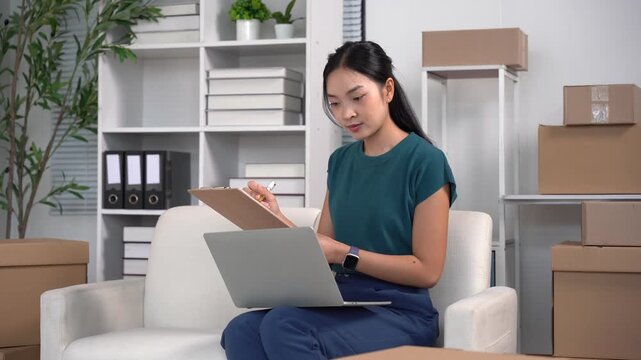 Entrepreneurial Spirit: A young female entrepreneur meticulously reviews documents on a clipboard, while a laptop rests on her lap, surrounded by an organized workspace with stacked boxes.