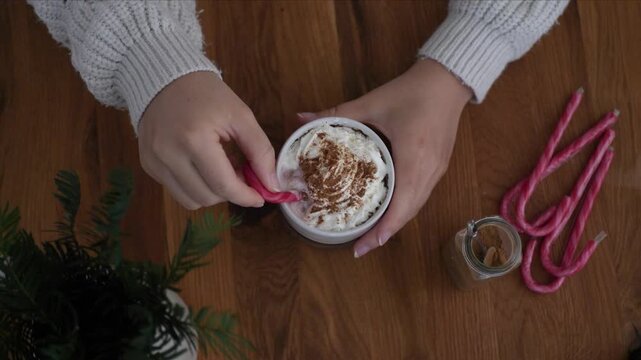 Hot chocolate with whipped cream stirred with candy cane spoon by woman hands, close up
