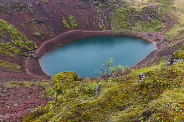 The color boundary along the inner crater wall appears at Kerid Crater, Iceland. Rain-darkened basin slopes lie in diffuse afternoon light above the crater lake during rainy overcast conditions. © Artem