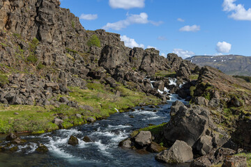 Fototapeta premium Several shallow drops through the rift channel appear along the Oxara River in Thingvellir National Park, Iceland. Water steps diagonally across basalt ledges beneath scattered clouds.