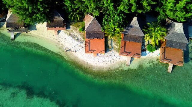 Aerial view of overwater bungalows with wooden decks and thatched roofs situated on a sandy beach along the turquoise coastline of Indonesia.