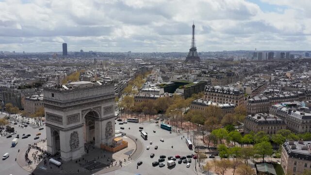 Aerial view of the Arc de Triomphe with the Eiffel Tower in the distance, surrounded by historic Parisian architecture and busy city streets in Paris, Ile-de-France, France.