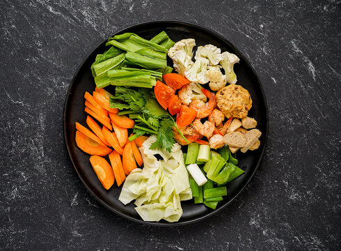 Top view of fresh raw chopped vegetables, shrimp, and meatballs prepared for cooking on a black plate over a dark stone background