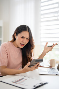 A woman is sitting at desk looking at calculator with a worried expression.