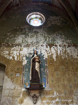 Statue of a saint holding roses and a cross beneath a stained-glass oculus with peeling fresco walls, Gers, Sarrant, France