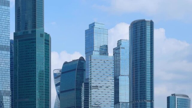 Timelapse looking up at the glass towers of the Moscow City business district. Cumulus clouds drift between the mirrored facades. Sunlight slides across the blue glazing high above the street.