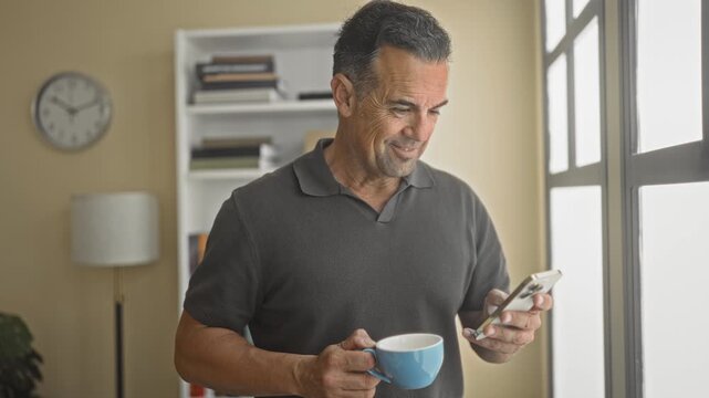 Man, middle aged, holding smartphone and blue coffee cup while looking at phone by window in building with bookshelf and clock visible; calm focus.